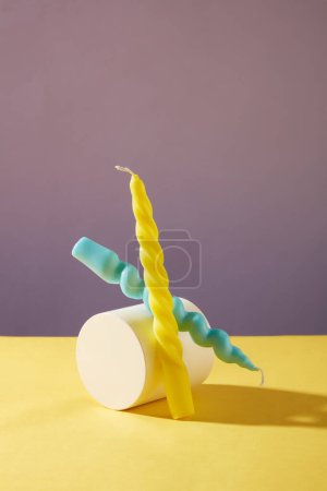 Photo for Two twisted candles in two colors: yellow and blue are placed next to a white podium on a yellow table with a purple background. Creative space for decoration. - Royalty Free Image