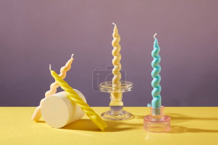 Photo for Two twisted candles are placed on glass candlesticks, two others are leaned next to the white podium. Yellow and purple background. Birthday with candles. - Royalty Free Image
