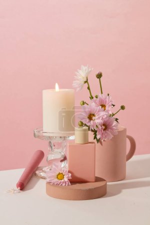 A burning candle is placed on a glass candlestick, an unlabeled perfume bottle is placed on a wooden podium and fresh flowers are on a pink and white background. Copy space for ads.