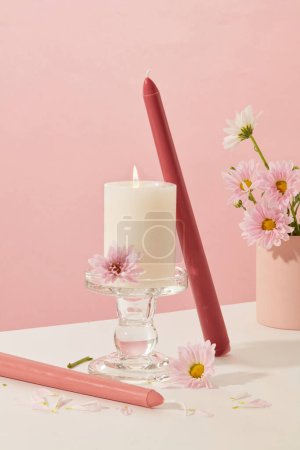 Photo for Front view of burning candle is placed on a glass candlestick. The surrounding is decorated with colored candles and fresh flowers on a white and pink background. - Royalty Free Image