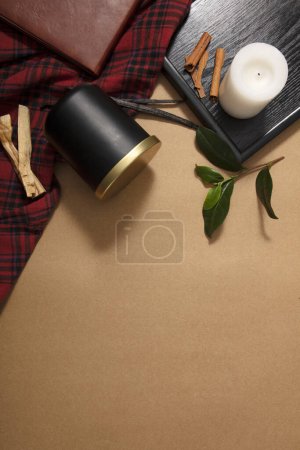 Téléchargez les photos : Un bocal de bougies parfumées noires est placé sur la table avec une nappe à carreaux, des feuilles vertes, un carnet, de la cannelle et un podium en bois sur un fond brun pastel. Espace vide pour le design. - en image libre de droit