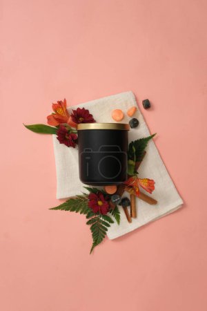 Téléchargez les photos : Un pot de bougies parfumées noires non étiquetées est placé à côté de fleurs fraîches, de feuilles vertes et de cannelle sur un fond rose avec un chiffon blanc. Maquette pour la publicité parfumée de bougie. - en image libre de droit
