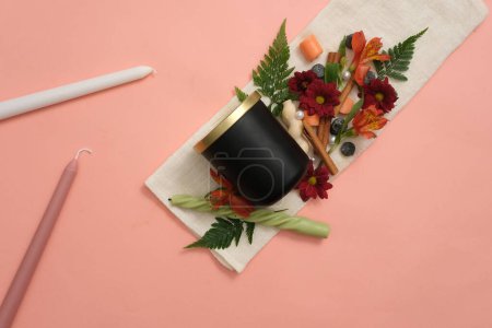 Téléchargez les photos : Bougies parfumées noires sans marque à côté de fleurs fraîches, bougies colorées, feuilles vertes et cannelle sur un fond rose avec un chiffon blanc. Modèle idéal pour la publicité de bougie parfumée. - en image libre de droit