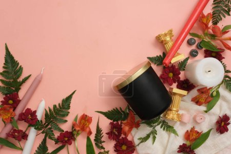 Téléchargez les photos : Pot de bougies parfumées non étiqueté avec des fleurs fraîches, des feuilles vertes et des bougies colorées sur un fond rose pastel. Beaucoup d'espace pour l'affichage des produits et la conception créative. - en image libre de droit