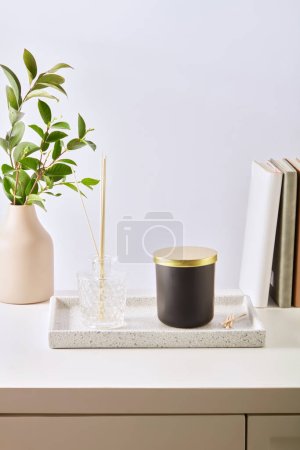 Photo for A jar of unlabeled scented candles and a bottle of essential oils are placed on the tray. Green leafy branches are placed in ceramic vases and books are on the table. White background for advertising. - Royalty Free Image