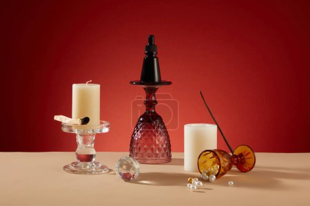 Photo for A black serum bottle sits on glass candlesticks amid decorative cups, white candles, and pearls on a beige table. Set against a red backdrop, a creatively inviting space for messaging. - Royalty Free Image