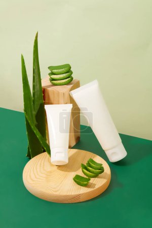 Photo for Front view of unbranded cosmetic tubes are displayed on a green table with a wooden platform and fresh aloe vera. Aloe vera effectively prevents and limits acne. - Royalty Free Image