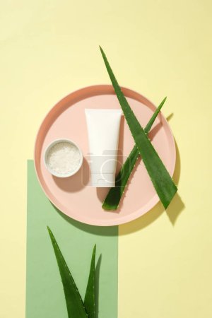 Photo for A pink ceramic plate is placed in the center of the frame, an unlabeled facial cleanser tube, a small bowl of salt and fresh aloe vera. Pastel background. Vegan cosmetics concept. - Royalty Free Image