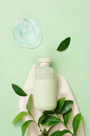Le thé vert est un ingrédient populaire dans les cosmétiques. Une bouteille de gel douche non étiquetée est placée à côté d'une serviette en tissu et de feuilles de thé vert frais, un verre d'eau sur un fond minimaliste.