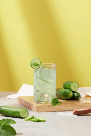 Photo for Yellow-white background with shadows. A glass of cucumber detox water and fresh cucumbers are placed on a wooden cutting board. The image brings a feeling of refreshment and freshness. - Royalty Free Image