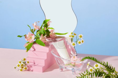 Téléchargez les photos : L'avant d'un tube cosmétique blanc non étiqueté est placé dans une mini baignoire en verre, à côté de laquelle se trouvent des fleurs fraîches et d'autres accessoires sur un fond pastel. Publicité cosmétiques naturels. - en image libre de droit