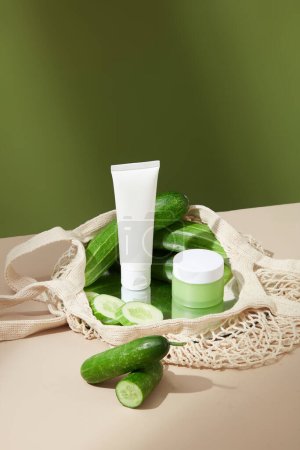 Photo for Close-up of a white tube and lotion jar on table with fresh cucumber. Mesh basket is displayed on beige-green moss background. Mockup cosmetic for advertising. - Royalty Free Image