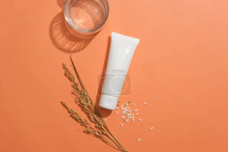 Photo for Centered unlabeled white tube with white rice and a glass on an orange background. Embracing a vegan cosmetics concept featuring rice extract. Ample copy space. - Royalty Free Image