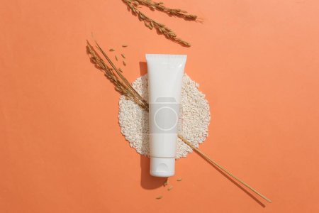 Photo for A tube without label placed on a pile of rice and decorated with wheat ears. Orange background. Empty label for cosmetic product mockup - Royalty Free Image