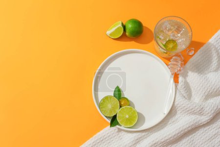 Foto de Vista desde arriba de limón, kumquat y hojas verdes decoradas en un plato redondo de cerámica, un vaso de agua lleno de hielo y un paño blanco. Espacio libre para publicidad. - Imagen libre de derechos