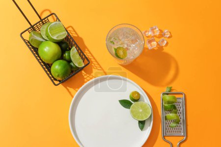 Photo for Lemon and kumquat are placed in an iron basket, a glass of water with ice, a grater with kumquat peel and a white ceramic plate on an orange background. Copy space for ads. - Royalty Free Image