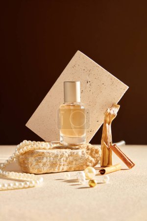 Photo for Scene for advertising perfume product with mockup for design. A glass bottle on block of stone decorated with pearl necklace, cinnamon sticks and white brick sheet on brown background. Front view - Royalty Free Image