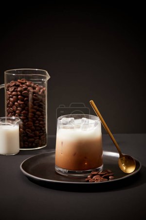 Téléchargez les photos : Vue de face d'un café au lait glacé placé sur un plateau avec une cuillère en métal. Café en grains et lait frais dans le bécher. Fond noir minimaliste pour la publicité produit. - en image libre de droit