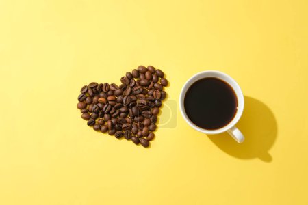 Téléchargez les photos : Grains de café disposés en forme de coeur et une tasse de café noir sur un fond jaune. L'application d'un masque à café aide à atténuer les taches de rousseur et à repousser les rides minuscules liées au vieillissement. - en image libre de droit