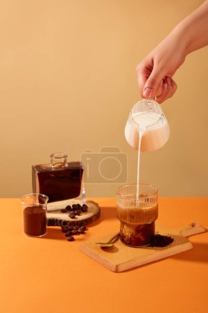 Téléchargez les photos : La main tient un verre de lait versé dans une tasse de café, entouré de grains de café, de poudre de café et de café noir. Le café aide à réduire le risque de maladie d'Alzheimer et de démence. - en image libre de droit