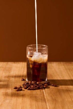 Photo for A stream of milk is pouring from above into a cup of black coffee on a wooden table with a brown background. Art space for food and beverage advertising. - Royalty Free Image