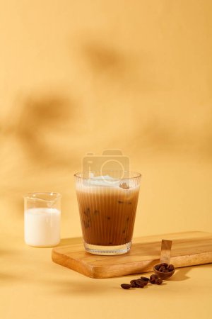 Foto de Vista frontal de una taza de café con leche se coloca en una plataforma de madera, junto a ella está la leche en un vaso de precipitados y granos de café sobre un fondo beige sombreado. Escena para publicidad de alimentos y bebidas. - Imagen libre de derechos