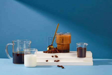 Photo for A glass of milky coffee is placed on a white platform with a metal spoon and coffee beans. Black coffee, fresh milk and coffee powder are stored in a beaker. Blue background for advertising. - Royalty Free Image