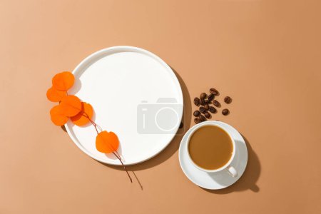 Téléchargez les photos : Une branche de feuille d'orange est placée sur une plaque de céramique blanche, une tasse de café au lait et quelques grains de café sur un fond brun. Espace vide pour l'affichage du produit. - en image libre de droit