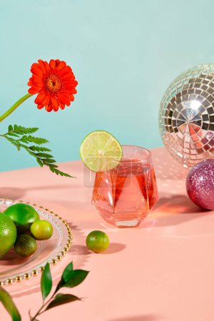 Téléchargez les photos : Sur une table, un verre contient un liquide rouge vif orné d'une tranche de citron, encadrée de fleurs fraîches et d'accessoires. Des teintes pastel de rose et de bleu créent un arrière-plan délicieux. - en image libre de droit