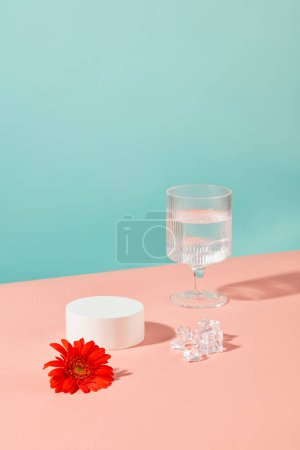 Téléchargez les photos : Un verre à vin contenant de l'eau, une fleur de gerbera rouge, de la glace et un podium blanc rond se détachent sur un fond bleu-rose. Afficher les produits sur un podium vide. - en image libre de droit