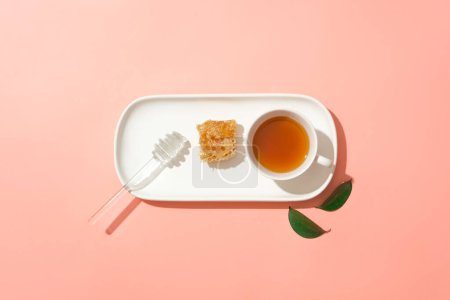 Photo for Minimal scene of a honey dripping, beeswax and a cup of tea arranged in a line on a dish. Pink background. Honey still contains high calories and sugar - Royalty Free Image