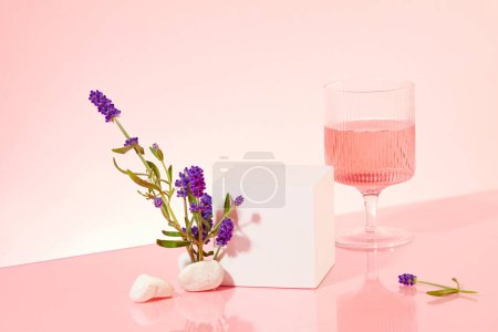 Téléchargez les photos : Sur un fond rose clair, un podium blanc et un verre à vin avec du liquide rose créent un affichage invitant, offrant amplement d'espace pour mettre en valeur la beauté dérivée de fleurs de lavande. - en image libre de droit
