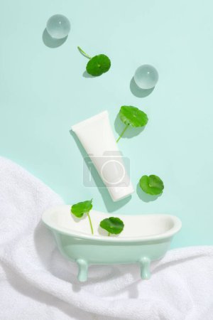 Photo for A tube of unlabeled lotion, two glass balls, a mini bathtub and pennywort leaves on pastel background. Gotu kola contains anti-inflammatory and antibacterial compounds. - Royalty Free Image