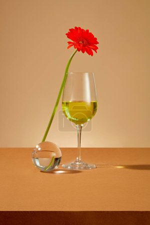 Téléchargez les photos : Un verre de vin rouge contenant du liquide jaune, une fleur de gerbera rouge et un globe de verre sont exposés sur une table brune. La beauté vient des fleurs fraîches. - en image libre de droit