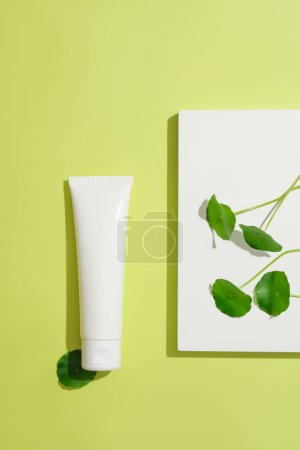 Photo for Close-up of an unbranded cosmetic tube, pennywort leaves on a white platform. Branding with blank labels. Cosmetic mockup for advertising. Top view. - Royalty Free Image