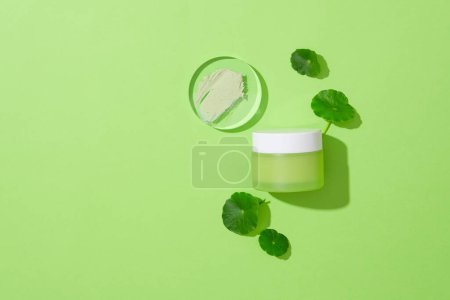 Photo for A smear of lotion on a glass platform, a jar of cosmetics and fresh pennywort displayed on a green background. Blank space for branding and advertising. - Royalty Free Image