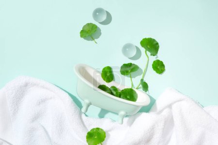 Photo for A towel, two glass marbles, a mini bathtub and fresh pennywort are displayed on a pastel background. Using products containing pennywort can help soothe the skin. - Royalty Free Image