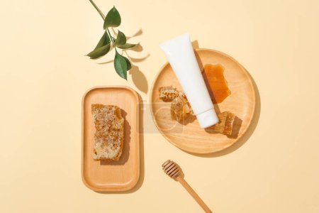 Téléchargez les photos : La cire d'abeille est stockée sur un plateau en bois, un tube cosmétique non étiqueté et du miel sur une assiette ronde en bois, une bruine de miel et des feuilles vertes sur un fond pastel. Espace idéal pour l'affichage des cosmétiques. - en image libre de droit