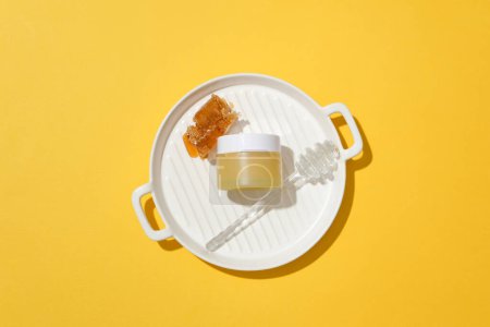 Téléchargez les photos : La cire d'abeille, un bocal cosmétique non étiqueté et une bruine de miel sont exposés sur un plateau rond en céramique. Miel contient un mélange de sucres et d'autres ingrédients nutritionnels. - en image libre de droit