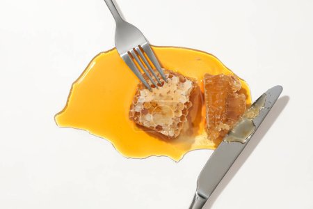 Téléchargez les photos : Une fourchette en métal est coincée dans de la cire d'abeille et un couteau repose dans du miel sur un fond blanc. La cire d'abeille contient des nutriments et des antioxydants, qui sont très bons pour le corps. - en image libre de droit