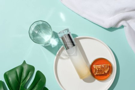 Photo for Honey and an unlabeled cosmetic bottle are displayed on a white ceramic plate, a glass of water, green leaves and a towel on a blue background. View from above. - Royalty Free Image