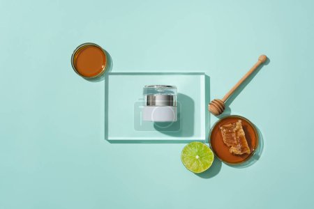 Photo for An unlabeled cosmetic jar is displayed on a glass podium, honey in a petri dish, fresh lemon and a honey drizzle on blue background. Exquisite space for cosmetic advertising. - Royalty Free Image