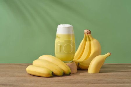 Foto de Un vaso de jugo de plátano colocado en un podio de madera, decorado con racimos de plátano. Los plátanos contienen una abundancia de vitamina A, que restaura la piel opaca y ayuda a iluminar la piel.. - Imagen libre de derechos