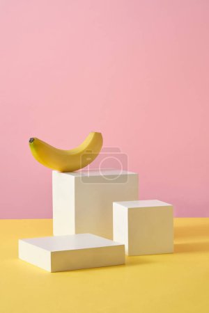 Quadratische Podeste in verschiedenen Größen werden mit einer Banane vor rosa Hintergrund dargestellt. Bananen (Musaceae) helfen abgestorbene Hautzellen zu entfernen, kontrollieren Öldrüsen und straffen die Poren.