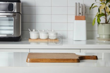 Selective focus, a wooden cutting board on a white tabletop, in the background is a blur kitchen interior - microwave, spice boxes, knife tray and a small potted plant. Scene for advertising products