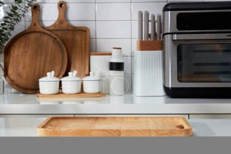 Modern kitchen interior with full appliances - wooden cutting board, spice boxes, knife tray and microwave on white tile wall. An empty wooden tray to place your product.