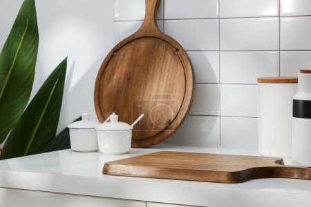 Some kitchen amenities decorated on white tile background - spice boxes, wooden cutting board and green leaves. Blank space for product presentation. Front view, menial scene for advertising
