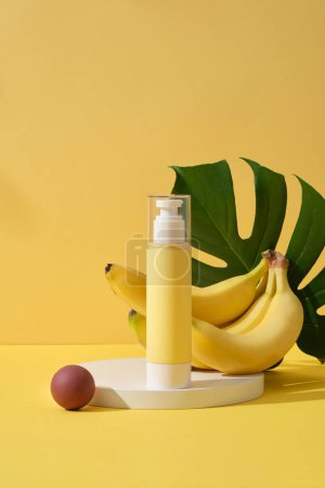 Scene for advertising and branding cosmetic with ingredient from banana - yellow bottle unlabeled on round podium with fresh ripe bananas and jungle monstera leaves on a yellow background