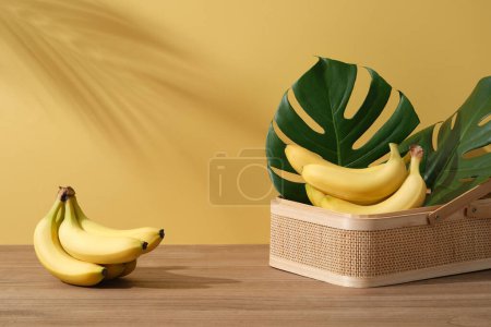 Background with minimalist style for product brand presentation and copy space - Fresh bananas on bamboo basket with monstera leaves on wooden table on yellow background with palm leaf shadow