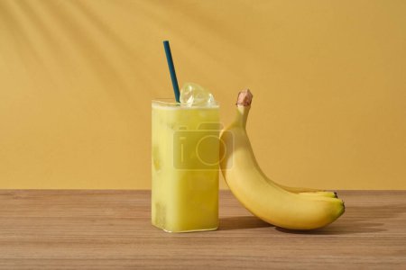 Front view of glass of banana juice and ripe yellow banana placed on wooden table on yellow background with palm leaf shadow. Scene for advertising product from banana with copy space
